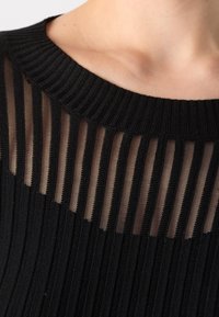 Gros plan d'un pull noir côtelé avec des rayures verticales transparentes près de l'encolure ronde laissant apparaître la peau nue en dessous.