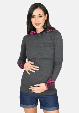 Schwangere Frau trägt langärmliges graues Oberteil mit pink gemusterten Bündchen und Kapuze, hält ihren Bauch und trägt dazu hochgekrempelte Jeansshorts.