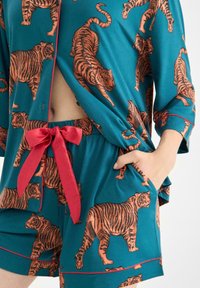 Conjunto de pijama de manga corta en color teal con un estampado de tigre en naranja, con un lazo rojo en la cintura y un bolsillo en los pantalones cortos.