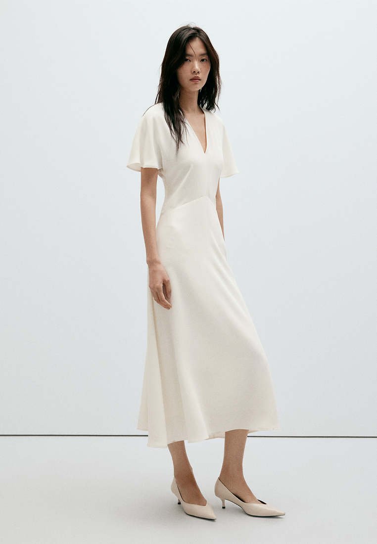 Femme portant une robe midi blanche avec des manches courtes volantées et un décolleté en V, associée à des chaussures beige à bout pointu et talons bas, debout devant un fond uni.