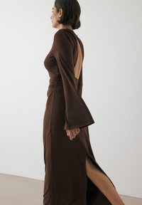 Abito marrone aderente con un'alta spacco laterale, maniche a campana lunghe e schiena scoperta, realizzato in tessuto liscio e elastico.