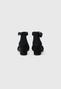 Schwarze Wildleder-Pumps mit Knöchelriemen, Blockabsatz, abgerundetem Zeh und silberner Schnallendetail am Riemen. Glatte Textur, minimalistisches Design.