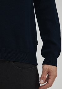 Pulover tricotat de culoare bleumarin, cu manșete și tiv cu nervuri, realizat dintr-un material moale. Etichetă mică de marcă vizibilă pe lateral. Purtați peste pantaloni negri.