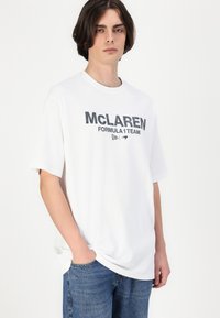 Balta kokvilnas T-krekls ar īsām piedurknēm, uz kura melnā krāsā ir druka "McLAREN FORMULA 1 TEAM". Valkāts ar ziliem džinsa bikšu.