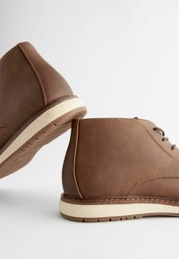 Botas de tobillo de cuero marrón con un acabado suave, puntera redondeada y suela de goma de color claro. Presentan detalles de costura y cordones.