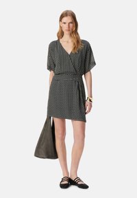 Vestido negro con estampado, escote en V, mangas cortas y cinturilla ajustada; combinado con un bolso de mano gris y zapatos planos negros.
