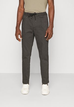 Homme portant un pantalon slim gris foncé avec cordon et des baskets blanches, debout devant un fond clair uni.