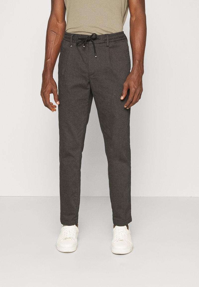 Homme portant un pantalon slim gris foncé avec cordon et des baskets blanches, debout devant un fond clair uni.