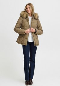 Khaki puffová bunda s kapucňou s kožušinovým lemom, prešívaný dizajn, predný zips a patentky, skombinovaná s tmavými džínsami a hnedými čižmami.