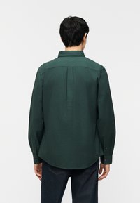 Άνδρας που  φοράει σκούρο πράσινο μακρυμάνικο πουκάμισο και σκούρο τζιν, όρθιος με την πλάτη προς το φακό, μπροστά σε απλό λευκό φόντο.