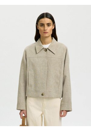 Frau mit langen dunklen Haaren, die eine beige, geknöpfte Jacke und helle Hose trägt und eine kleine braune Tasche vor neutralem Hintergrund hält.