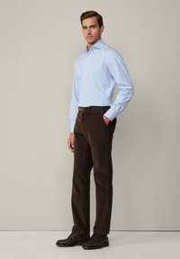 Chemise bleu clair à manches longues, associée à un pantalon en velours côtelé marron. Un homme se tient de manière confiante, les mains dans les poches, portant des chaussures noires.