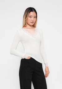 Jeune femme aux longs cheveux portant un haut à manches longues à col en V bordé de dentelle blanche et un pantalon noir à motifs, une main dans la poche.