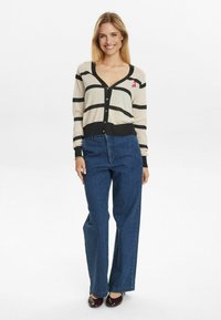 Striped cardigan in cream and black, V-neck, button front, with pink cherry detail. Paired with wide-leg blue jeans and dark shoes.