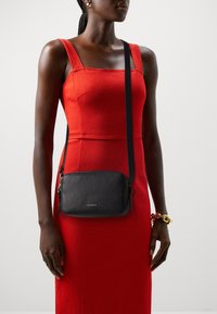 Borsa a tracolla in pelle nera texture con chiusura a zip e tracolla regolabile, indossata sopra un vestito rosso aderente. Dettaglio del logo dorato sulla parte anteriore.