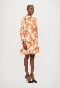 Blumenmusterkleid in Creme mit orangefarbenen Mustern, mit hohem Kragen, gerafften Ärmeln und einem gestuften Rock. Schwarze High-Heels.