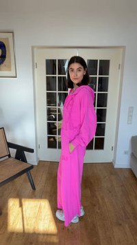 Žena stojící uvnitř, oblečená v jasně růžové mikině s kapucí a odpovídajících kalhotách s širokými nohavicemi, v bílých teniskách, vedle židle a skleněných dveří.