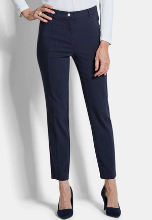 Femme portant un pantalon cigarette marine ajusté à la cheville, un haut blanc à manches longues et des chaussures à talons hauts marine, debout devant un fond clair.