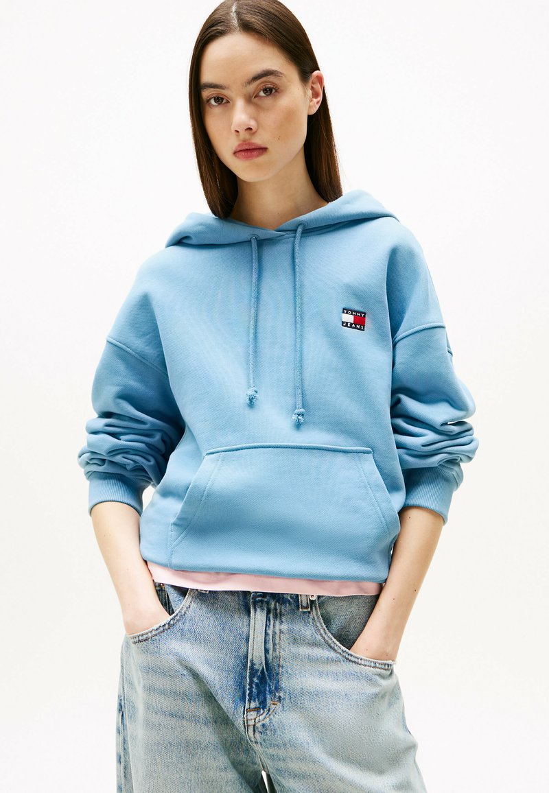 Sudadera de color azul claro con mangas largas, capucha con cordón y bolsillo frontal. Presenta un pequeño logo rojo y azul en el pecho.