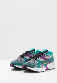 Sportskor med övre del av nät i svart, teal och lila med ett strukturerat mönster, en vit mellansula och en lila yttersula.