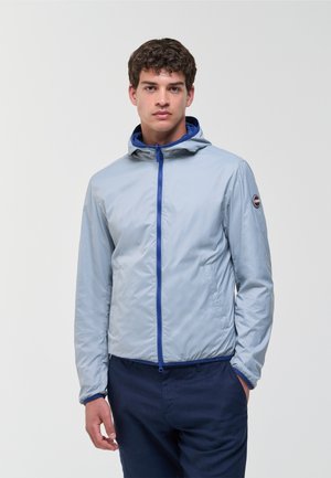 Junger Mann trägt eine hellgraue Kapuzenjacke mit blauem Reißverschluss und marineblaue Hose, steht vor einem schlichten hellen Hintergrund.