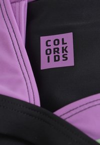 Lila och svart tyg med kontrasterande sömmar; har en rektangulär lila etikett med präglad svart text "COLOR KIDS."