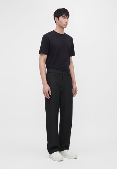 Homme debout portant un t-shirt noir, un pantalon noir et des baskets blanches sur un fond uni gris clair.
