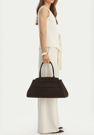 Žena v bílém tílku a širokých kalhotách držící velkou černou kabelku za rukojeti na bílém pozadí.