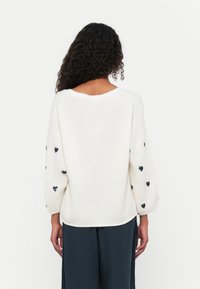 Maglione bianco lavorato a maglia con vestibilità ampia, scollo rotondo e maniche a sbuffo 3/4. Decorato con piccoli motivi a forma di cuore nero sulle maniche.