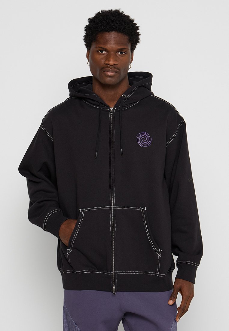Sudadera con cremallera negra, con bolsillos frontales, costuras blancas y un logo en espiral morado en el pecho. Fabricada con una tela suave y texturizada.