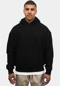 Sudadera negra con capucha y un bolsillo frontal, con el texto "Drops" en una impresión sutil en tonos oscuros. Material de algodón con un corte relajado.