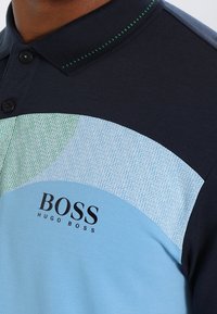 Mörkblå polo skjorta med ljusblå, grön och vit grafisk panel, svarta knappar och "BOSS HUGO BOSS"-logotyp på bröstet.
