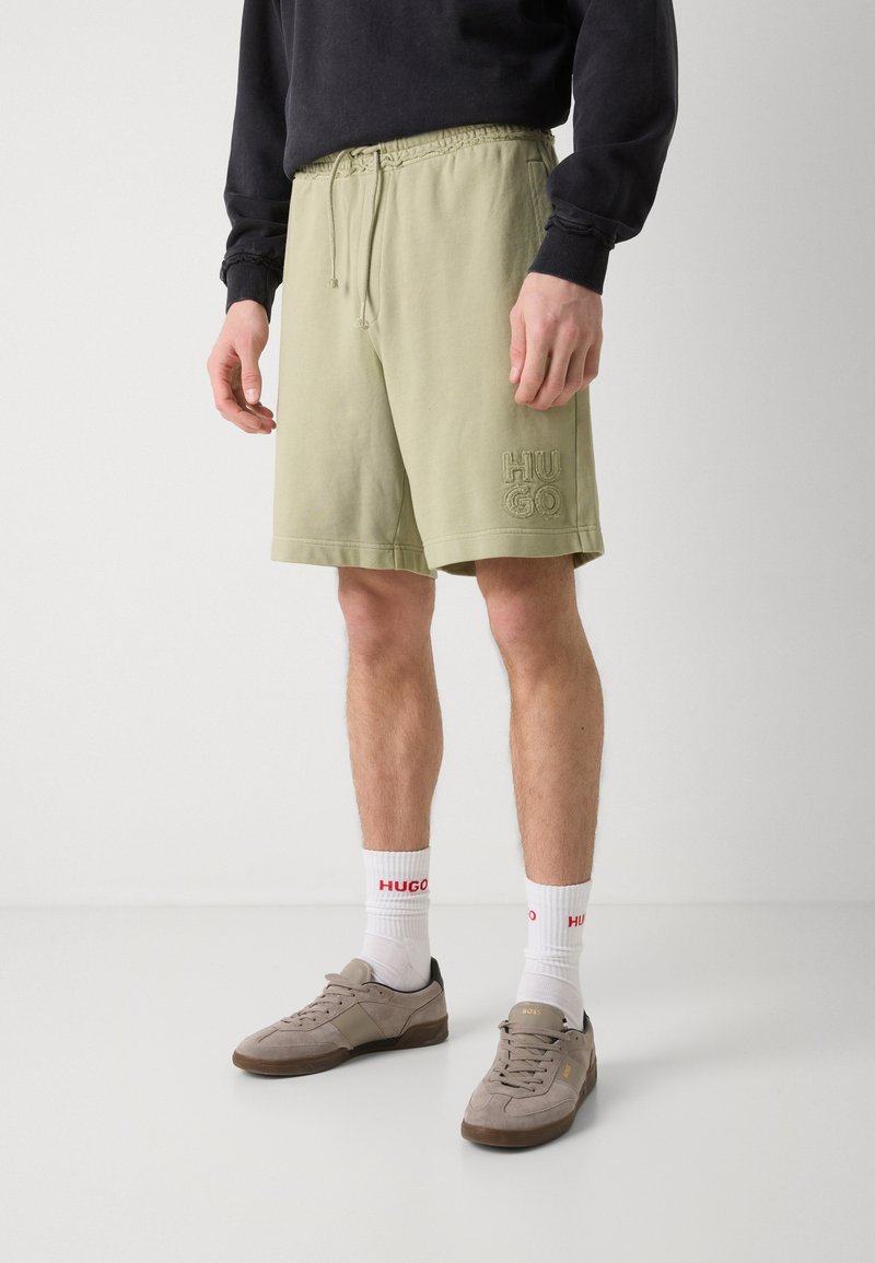 Hellgrüne Shorts mit elastischem Taillenbund, verziert mit gesticktem "HUGO"-Logo. Kombiniert mit grauen Turnschuhen und weißen Socken mit roten Akzenten.