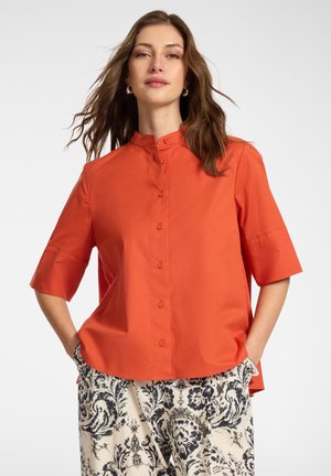 Kobieta ubrana w jaskrawą pomarańczową koszulę na guziki z kołnierzykiem mandarynka oraz czarno-białe spodnie w wzory, stoi z rękami w kieszeniach.