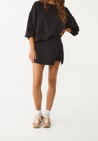 Sudadera negra corta con mangas largas acompañada de una falda envolvente negra. Zapatillas beige con detalles completan el look.