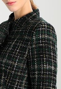 Donna che indossa un cappotto a quadri tessuto con un alto colletto, di colore verde scuro e nero, mostrando una vista parziale del collo e delle labbra.
