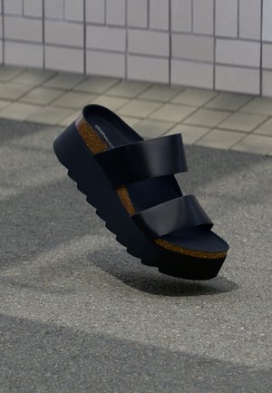 Μαύρο πλατφόρμα σανδάλι με χοντρή σόλα από φελλό και δύο φαρδιές λουρίδες, τοποθετημένο σε υφή δαπέδου κοντά σε πλακιδωτό τοίχο.