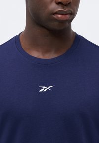 Gros plan d'une personne portant un t-shirt uni bleu marine avec un petit logo blanc Reebok centré sur la poitrine.