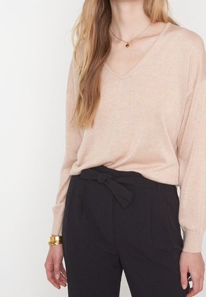 Femme portant un pull beige clair à col en V, un pantalon noir taille haute avec ceinture nouée, un bracelet en or et un petit collier pendentif.