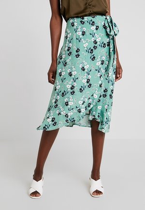 Kobieta w zielonej kwiecistej spódnicy kopertowej z wiązaniem na boku, w białych sandałach na obcasie z odkrytymi palcami i brązowej bluzce, stojąca na jednolitym tle.