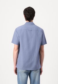 Kortermet skjorte i lyseblått stoff, har krage og bakstykke, med en avslappet passform og lett buet hem. Denim jeans synlige.