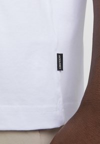 Bijela pamučna majica s okruglim izrezom i malom crnom etiketom s natpisom "JACK&JONES" na bočnoj šav. Glatka tekstura, kratki rukavi.