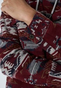 Rød og grå mønstret stofjakke med en knapmanchet, der har geometriske designs og en blød tekstur.