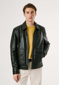 Chaqueta de cuero negra con cuello, cremallera frontal, dos bolsillos en el pecho y una textura suave, llevada sobre un suéter amarillo.