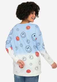 Camisa de manga larga azul claro con gráficos de caras sonrientes dispersos en negro y rojo, que se desvanecen a blanco en el dobladillo. Tejido suave, corte relajado.