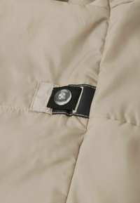 Beige vadderad tyg med en slät yta, prydd med ett svart band och en genomskinlig knapp, säkrad med kontrasterande sömnad.