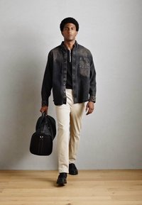 Black denim shirt, black polo, beige pants, black shoes, holding a black bag, standing on wooden floor against a gray background.