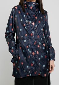 Giacca impermeabile blu navy con motivo floreale in tonalità di rosso e blu, caratterizzata da vita aderente, polsini elasticizzati e cappuccio.