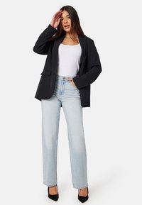 Zwarte blazer met geknipte revers, witte tanktop, lichtblauwe wijde jeans en zwarte hoge hakken. De blazer heeft voorzakken.