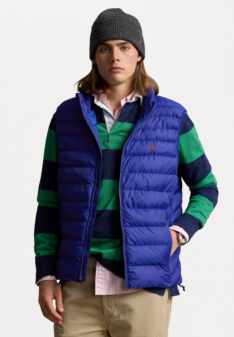 Blaues Steppweste mit einem gesteppten Design, getragen über einem grün-blauen gestreiften Langarmshirt. Zubehör umfasst eine dunkle Mütze und eine Uhr.